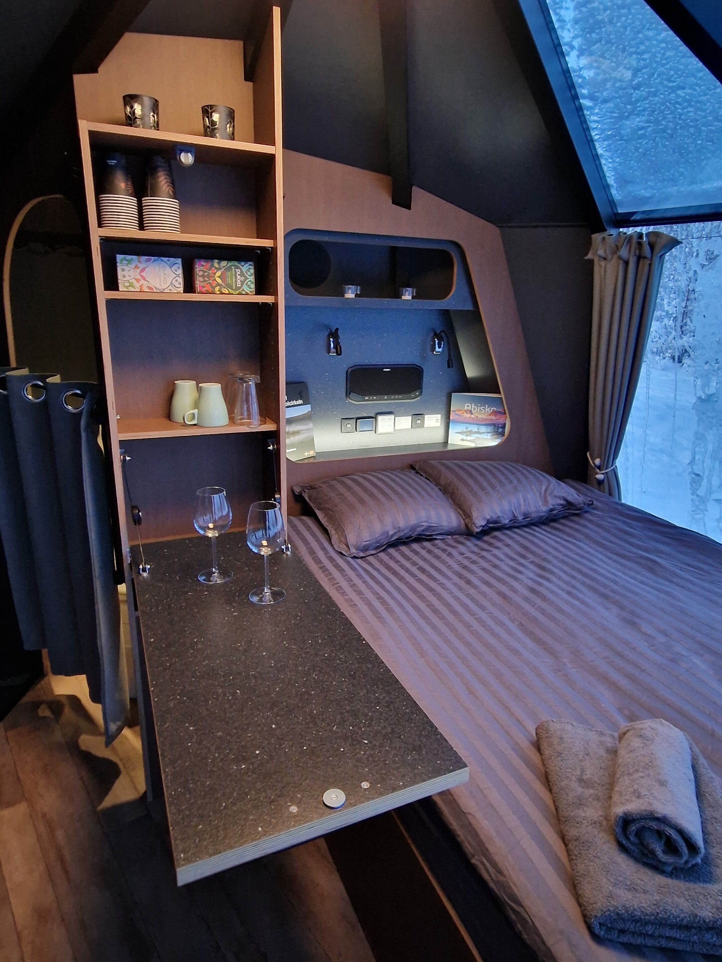 Un dormitorio acogedor con cama, una mesita con copas de vino, estantes con objetos decorativos y una ventana con vistas al cielo.