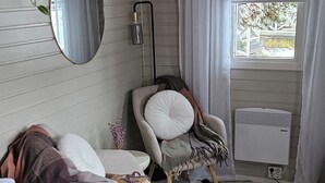Una habitación con pared de listones de madera, un espejo redondo, una silla con cojín blanco, un pequeño calentador y una ventana con cortinas.