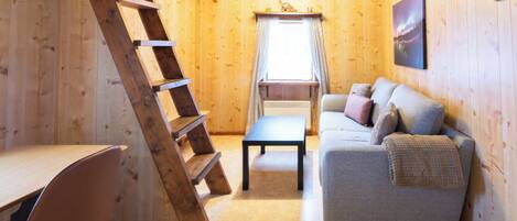 A wooden interior with a sofa, a table, a ladder, and a window with curtains.