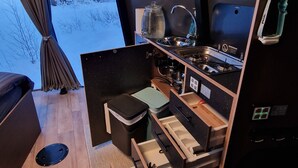 Cocina compacta con estufa, pileta y cajones de almacenamiento. Una ventana muestra un paisaje nevado al exterior.