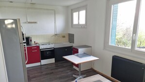 Cocina compacta con refrigerador, estufa, microondas y pileta. Hay una mesita con una botella y cenicero. La habitación tiene una ventana y un radiador.