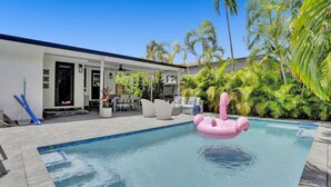 A swimming pool with a pink flamingo float, surrounded by palm trees and a covered patio area with outdoor furniture. - Casa Coral with  Pool & Charcoal BBQ area (Miami)