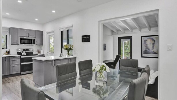 A modern kitchen with stainless steel appliances, a dining area with a glass table and grey chairs, and a living space with a sofa and artwork. - Casa Coral with  Pool & Charcoal BBQ area (Miami)