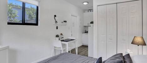 A compact bedroom with a bed, bedside table, and a desk with a chair. There is a window providing natural light and a view of the outside.