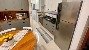 A compact kitchen with a refrigerator, stove, and white cabinets. There is a wooden dining table with a lace table runner and a vase of yellow flowers. - Apê Harmonia  (Ubatuba)