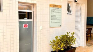 A building exterior with tiled walls, a door with a red sticker, a signboard, and a potted plant. - Apê Harmonia  (Ubatuba)