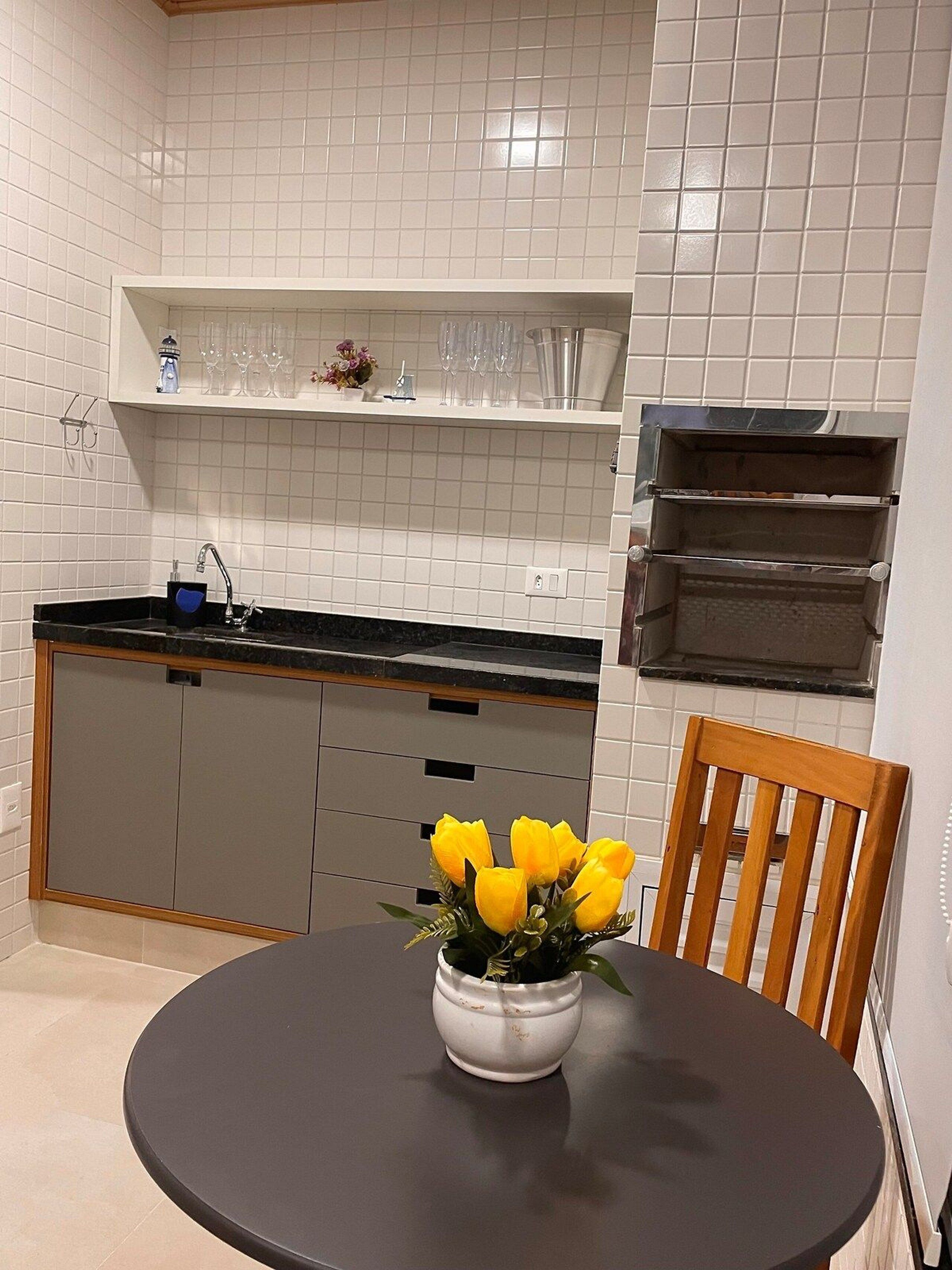 A kitchen with white tiled walls, a black countertop, a sink, a built-in oven, and a dining area with a round table and a chair.