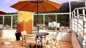 A terrace with a tiled floor, a table with chairs, a large umbrella, and a view of buildings and hills in the background. - Apê Harmonia  (Ubatuba)