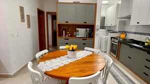 A dining area with a wooden table and white chairs, a kitchen with white cabinets, and a small sailboat decoration on the ceiling. - Apê Harmonia  (Ubatuba)