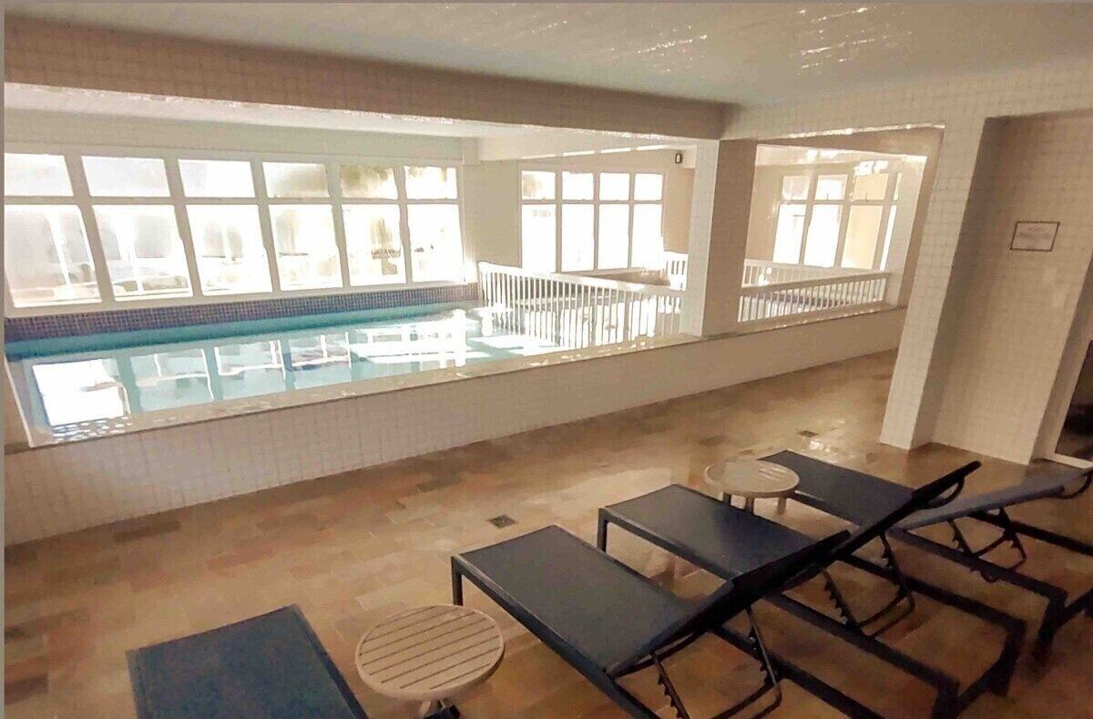 Ein Innenpoolbereich mit Blick auf den Pool durch große Fenster, Liegestühle und ein kleiner runder Tisch.