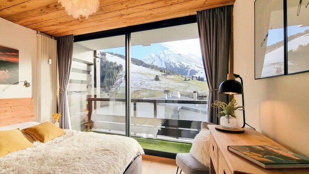 Un dormitorio con una cama grande, un escritorio con lámpara y vistas a un paisaje nevado de montañas a través de un ventanal.