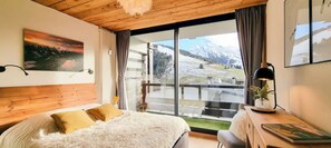 A bedroom with a large bed, a desk with a lamp, and a view of a snowy mountain landscape through a large window. - The White Bear - Spacious and Comfortable Apartment (La Clusaz)