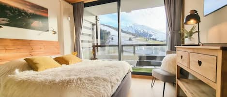 Ein Schlafzimmer mit einem großen Bett, einer hölzernen Kommode und Blick auf Berge durch ein großes Fenster.