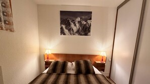 A hotel room with a bed, two bedside lamps, a mountain view painting, and a folded towel on the bed. - Apartment of 30m2 with 1 room, 1 bunk bed area and 1 living room with sofa (Les Allues)
