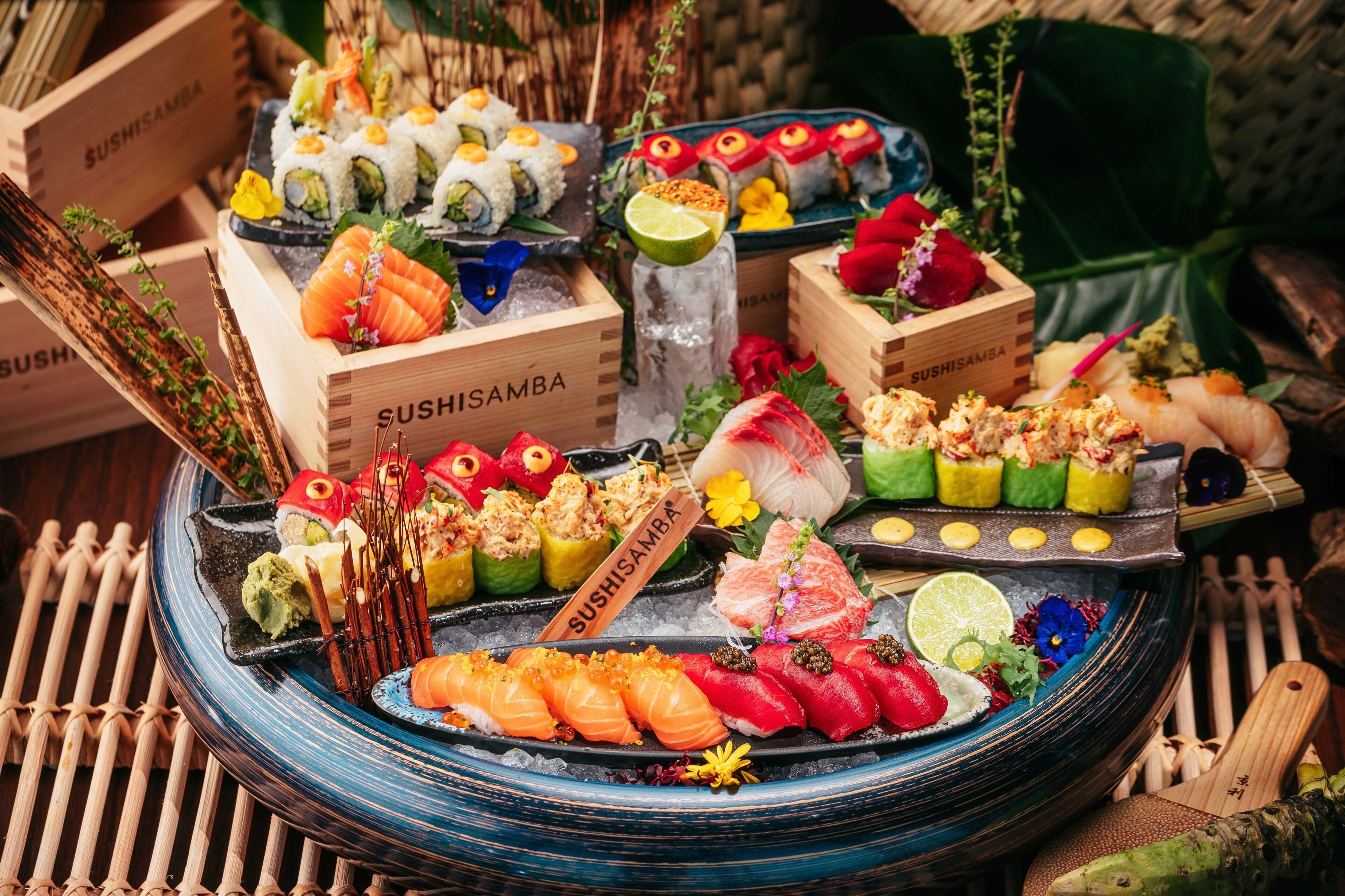 A variety of sushi dishes displayed on a round table with ice, surrounded by wooden boxes labeled "SUSHISAMBA".
