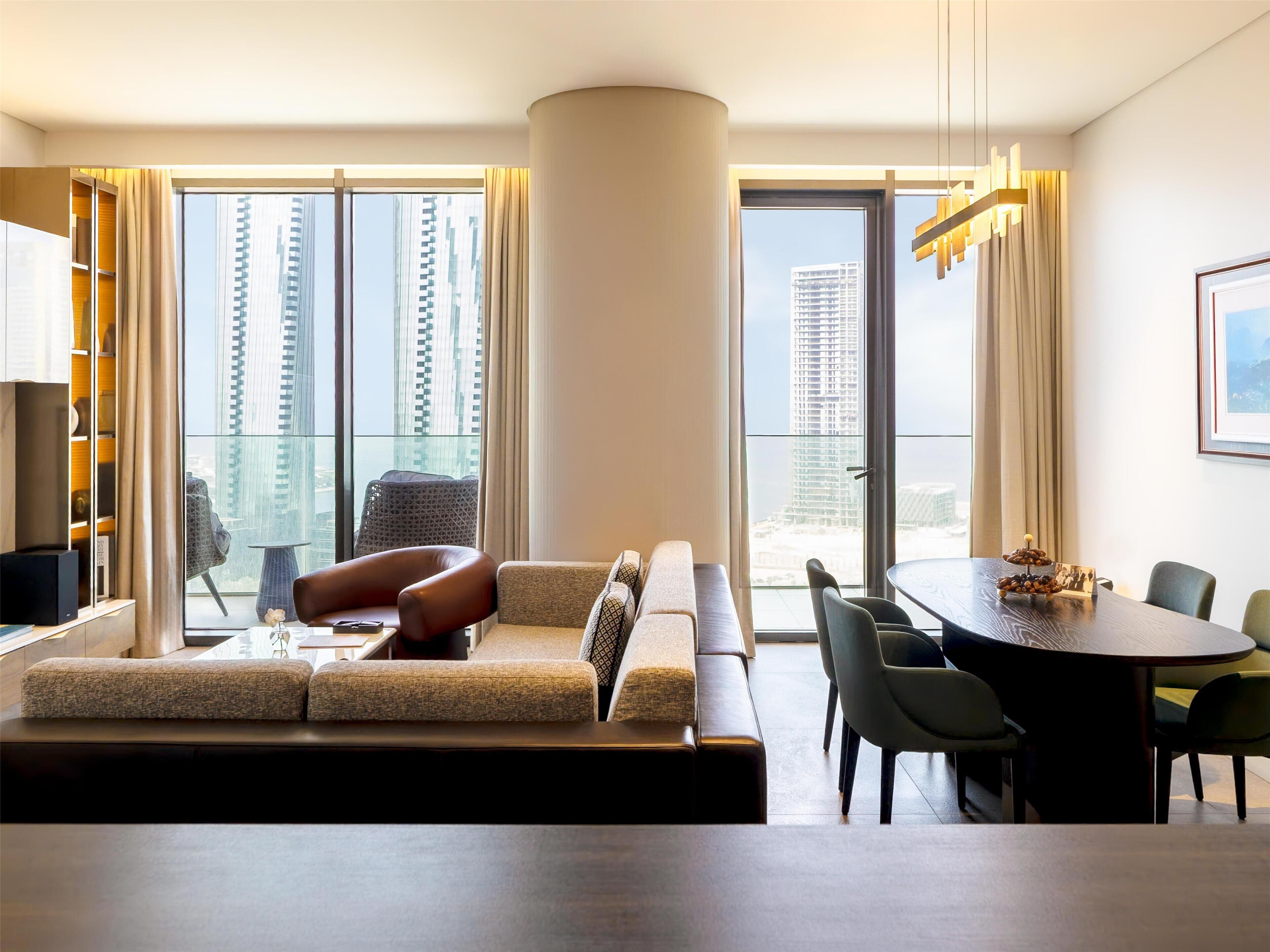 A modern living room with a sofa, dining table, and chairs. Large windows offer views of tall buildings.