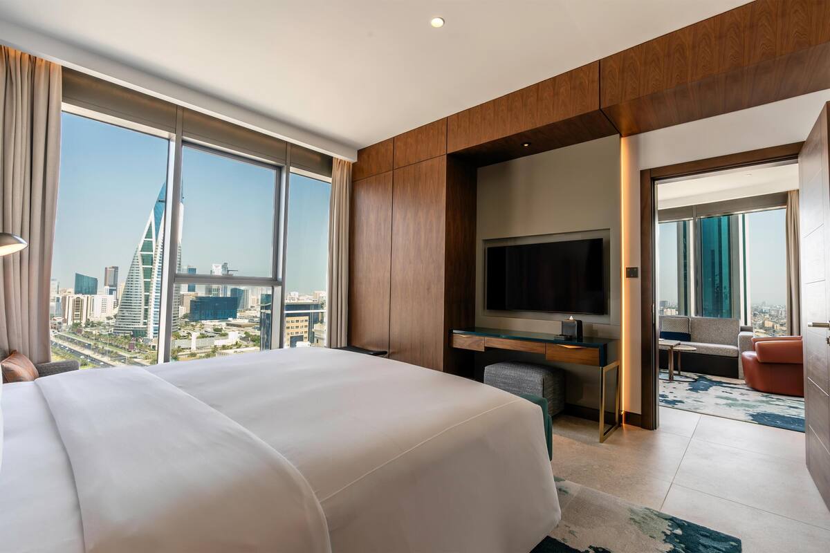 A modern hotel room with a large bed, a desk with a flat-screen TV, and a city view.