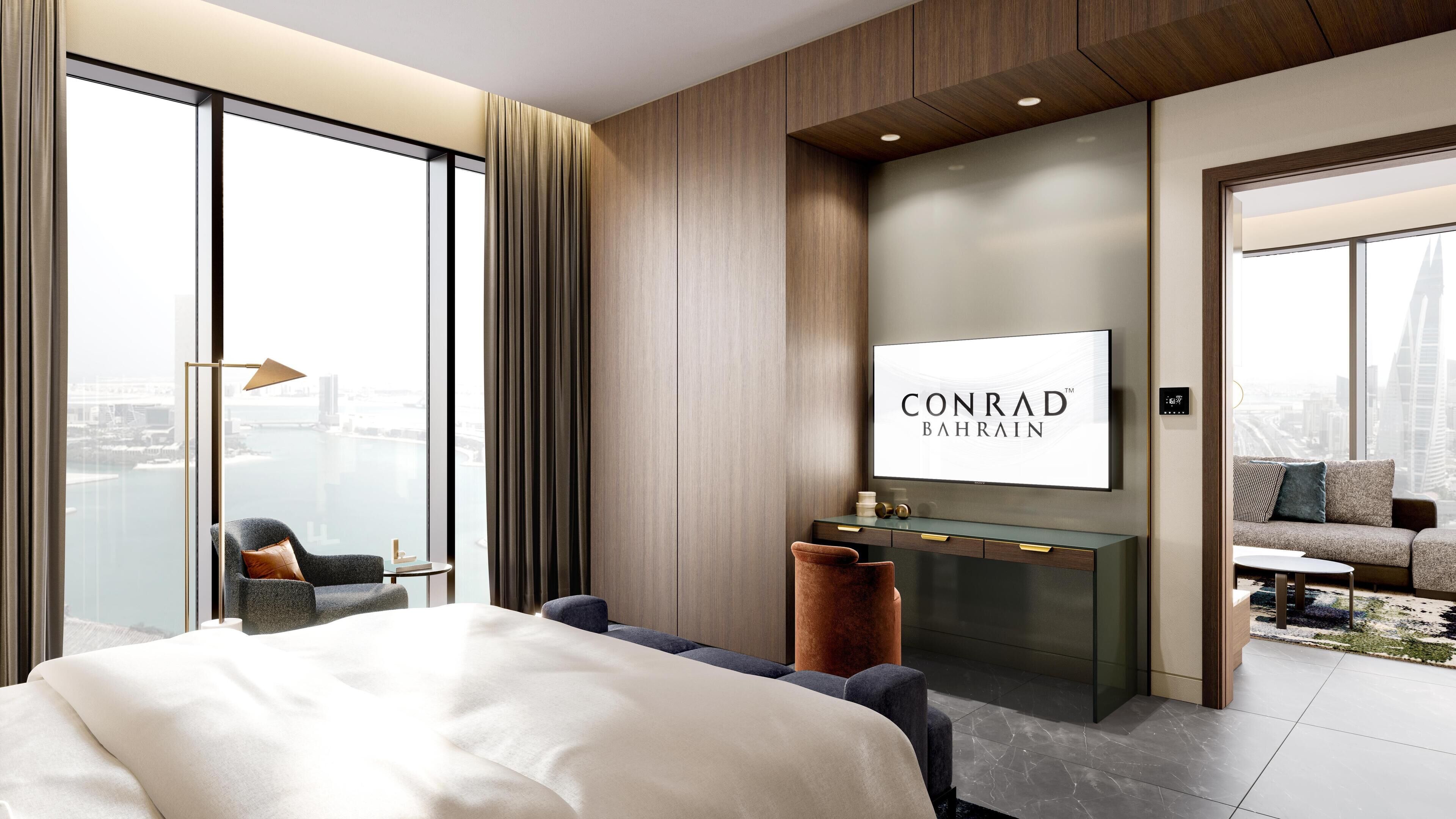 A modern hotel room with a large bed, a seating area, a TV, and a view of the city.