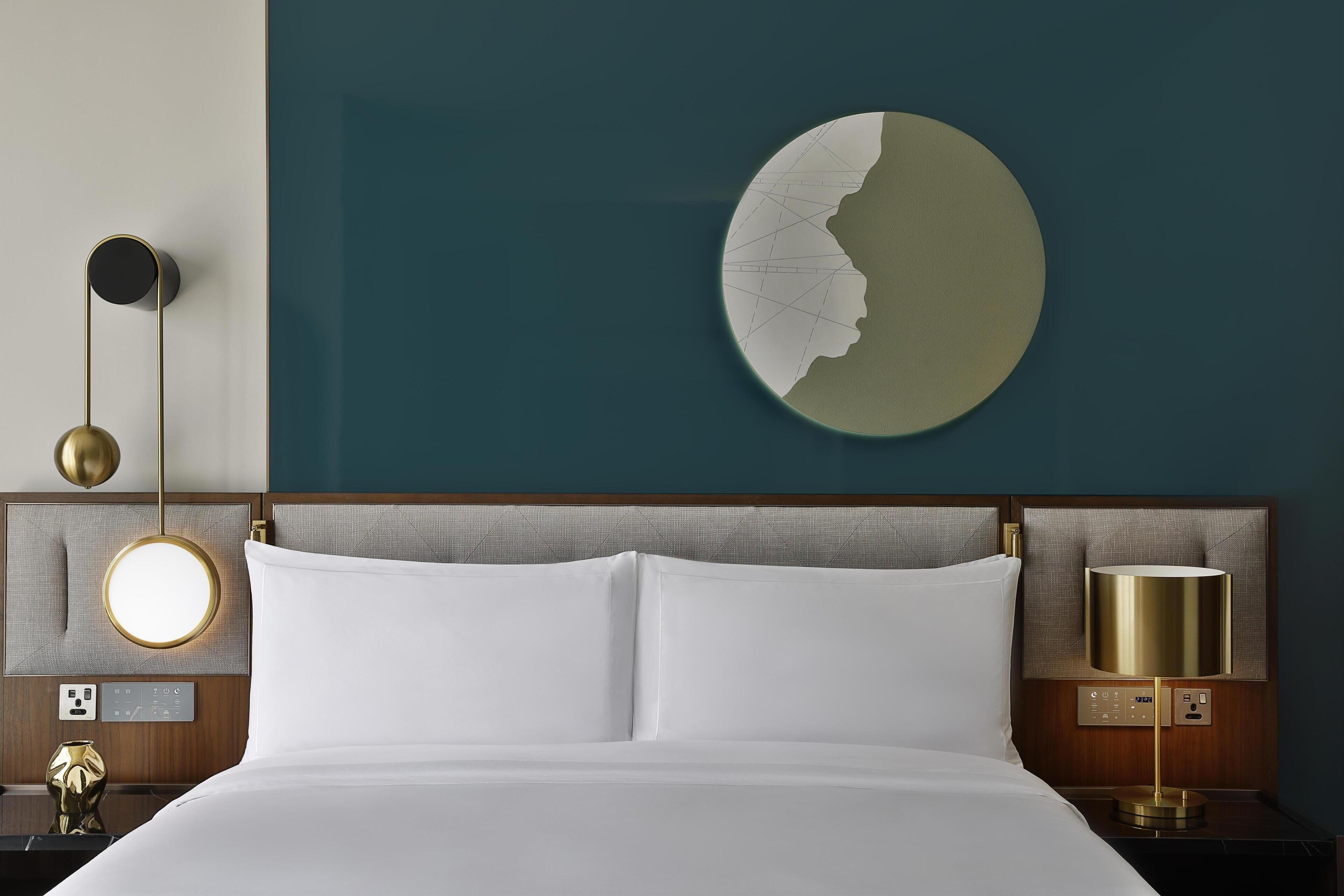 A hotel room with a neatly made bed, a wall-mounted round mirror, a wall clock, and a modern lamp.