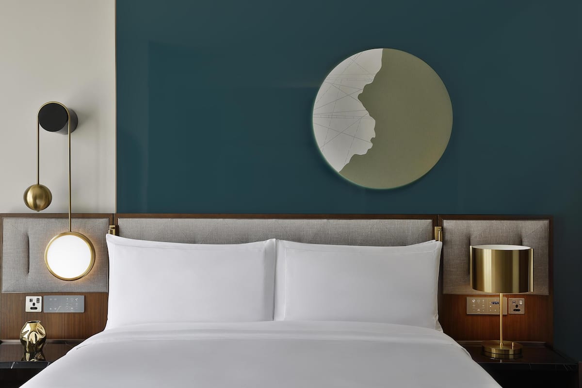 A hotel room with a neatly made bed, a wall-mounted round mirror, a wall clock, and a modern lamp.