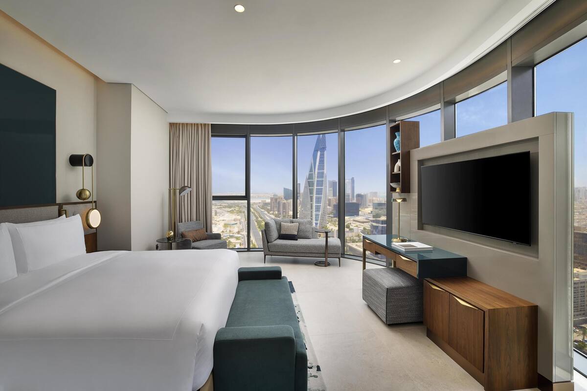 A modern hotel room with a large bed, a seating area with a view of the city, a flat-screen TV, and a wooden cabinet.