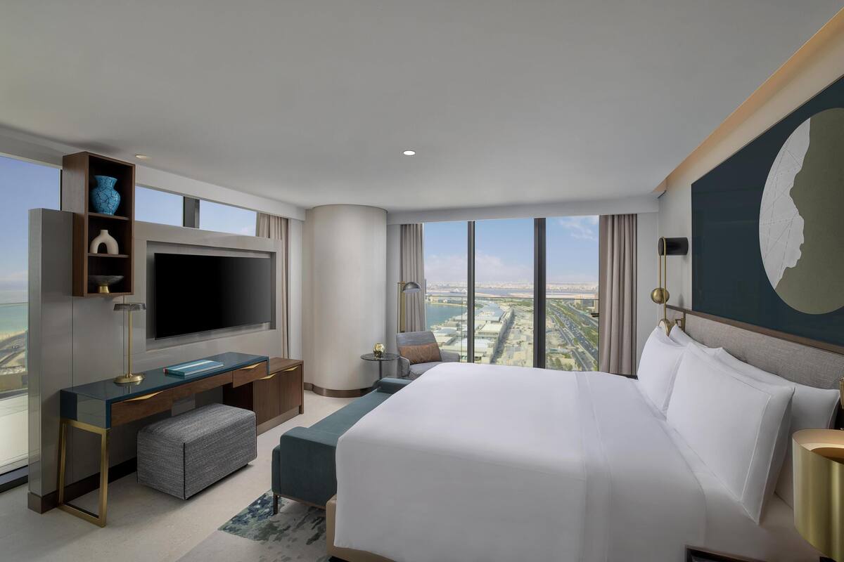A modern hotel room with a large bed, a flat-screen TV, a desk with a chair, and a view of the cityscape.