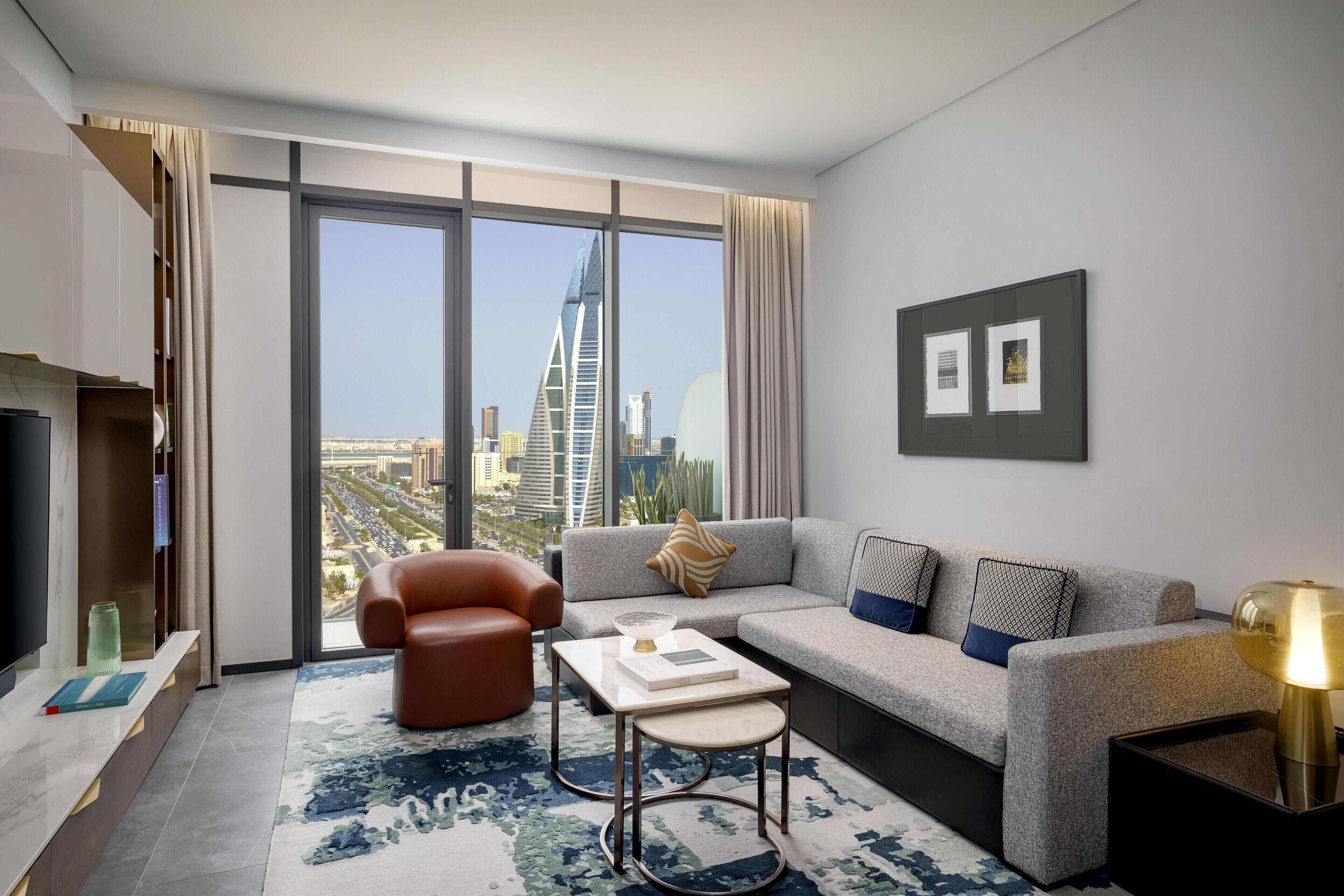 A modern living room with a sofa, armchair, and coffee table, large windows with a city view, and a flat-screen TV.
