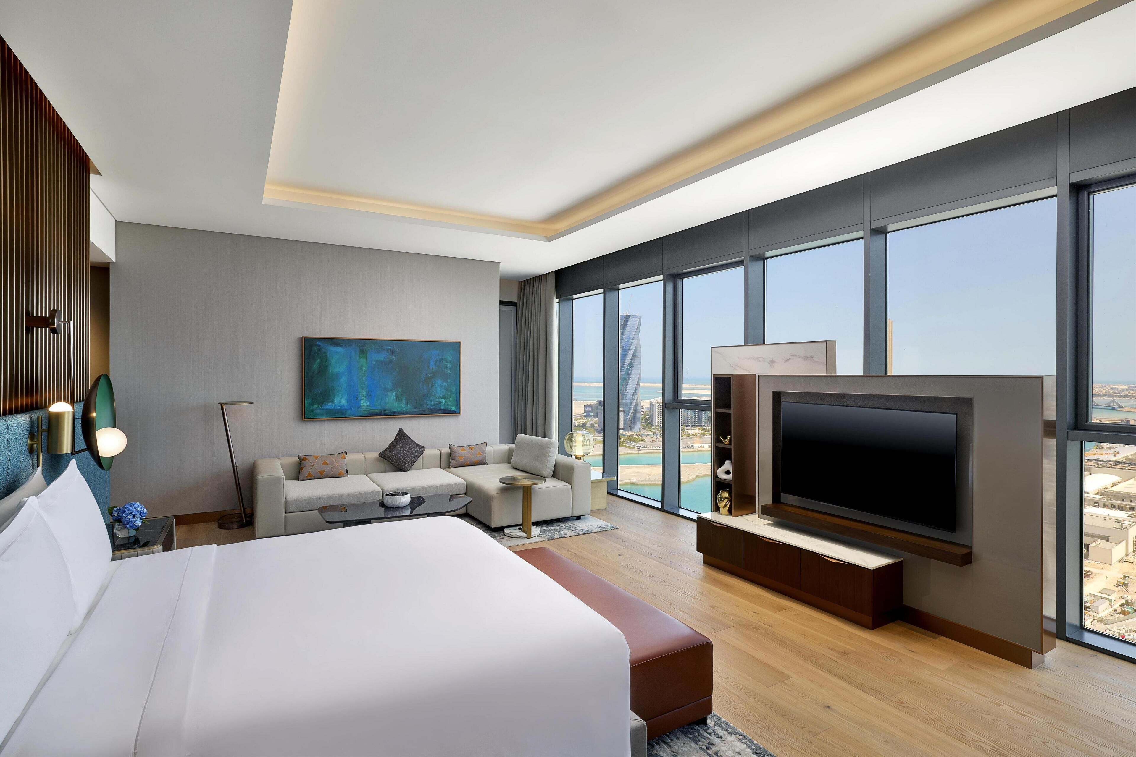 A modern hotel room with a large bed, a sofa, a TV, and a city view.