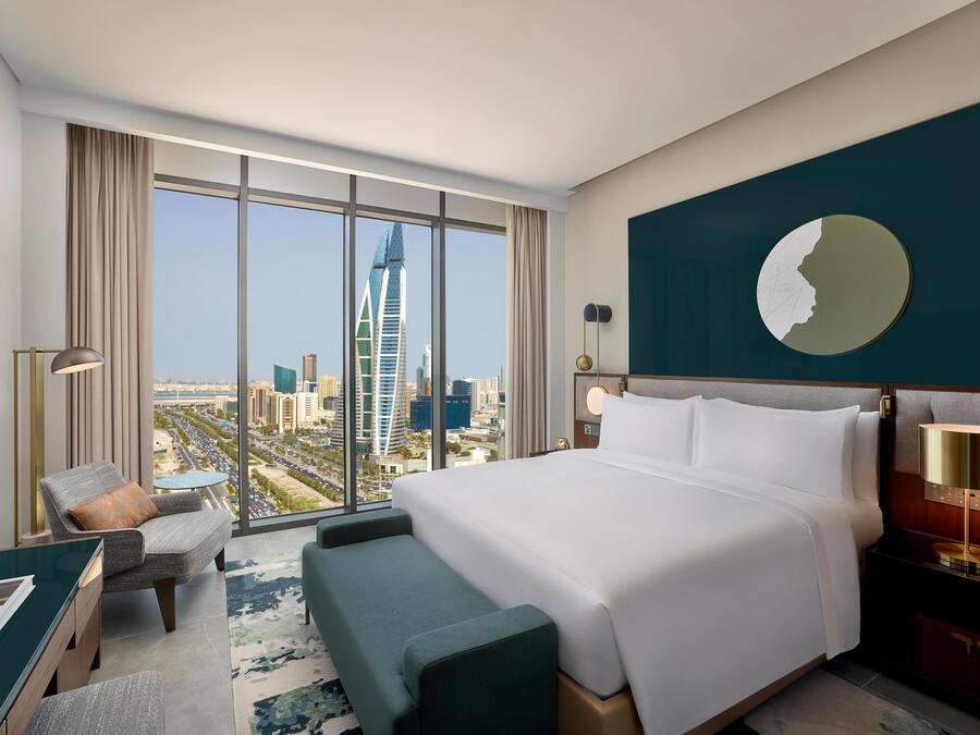 A modern hotel room with a large bed, a desk, a chair, and a view of a cityscape including a tall building.