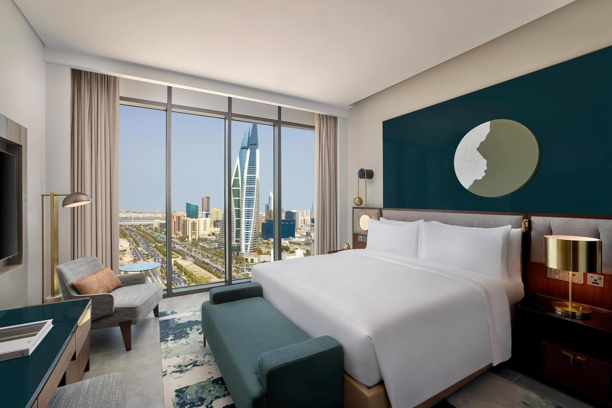 A hotel room with a large bed, a desk, a chair, and a view of a cityscape including a tall building.
