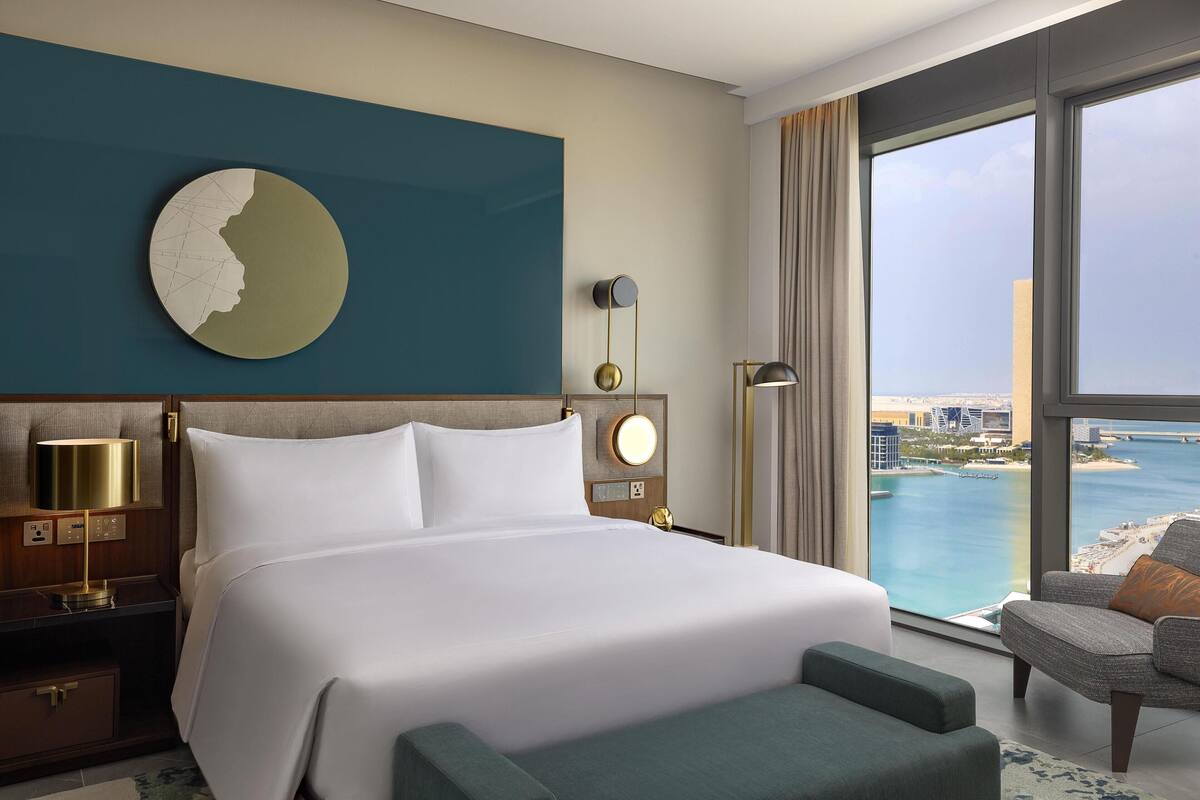 A hotel room with a large bed, a view of the sea, a round wall decoration, and a modern lamp.