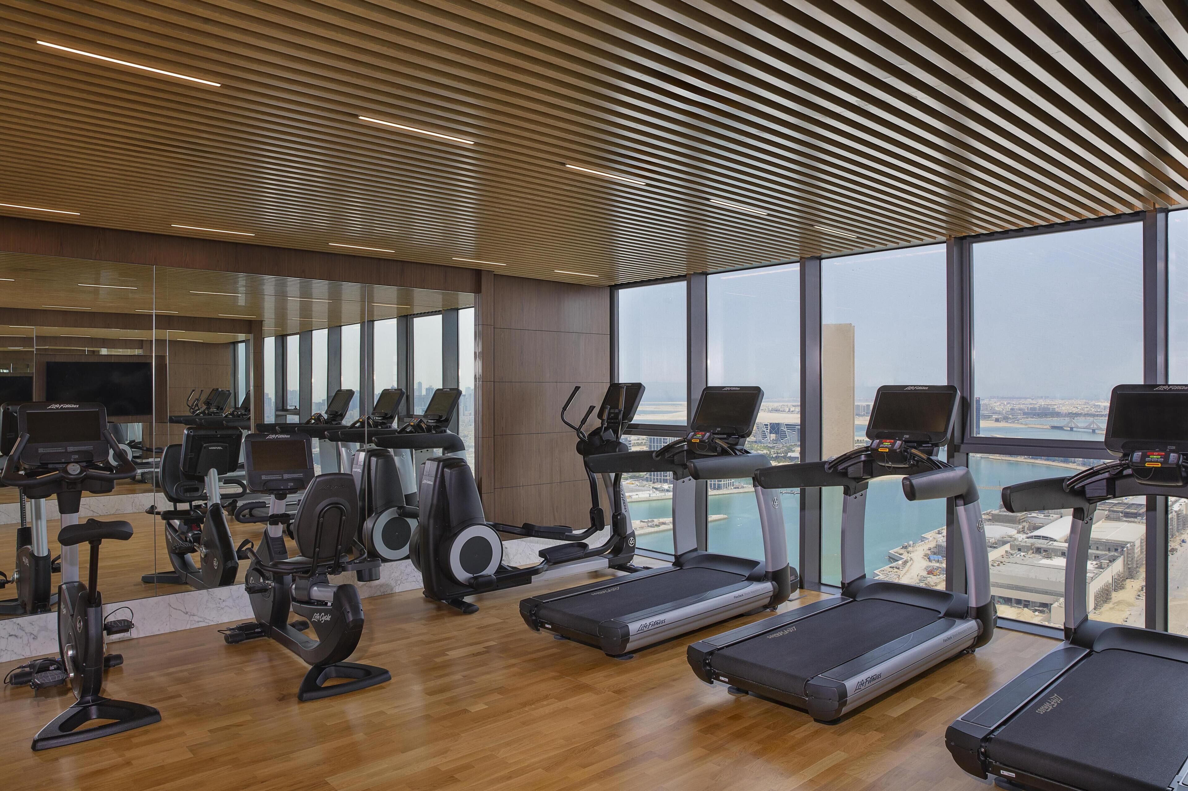 A modern gym with various cardio machines, large windows, and a city view.