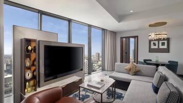 Habitación de hotel moderna con televisor grande, asientos cómodos, zona de comedor y vistas a la ciudad.