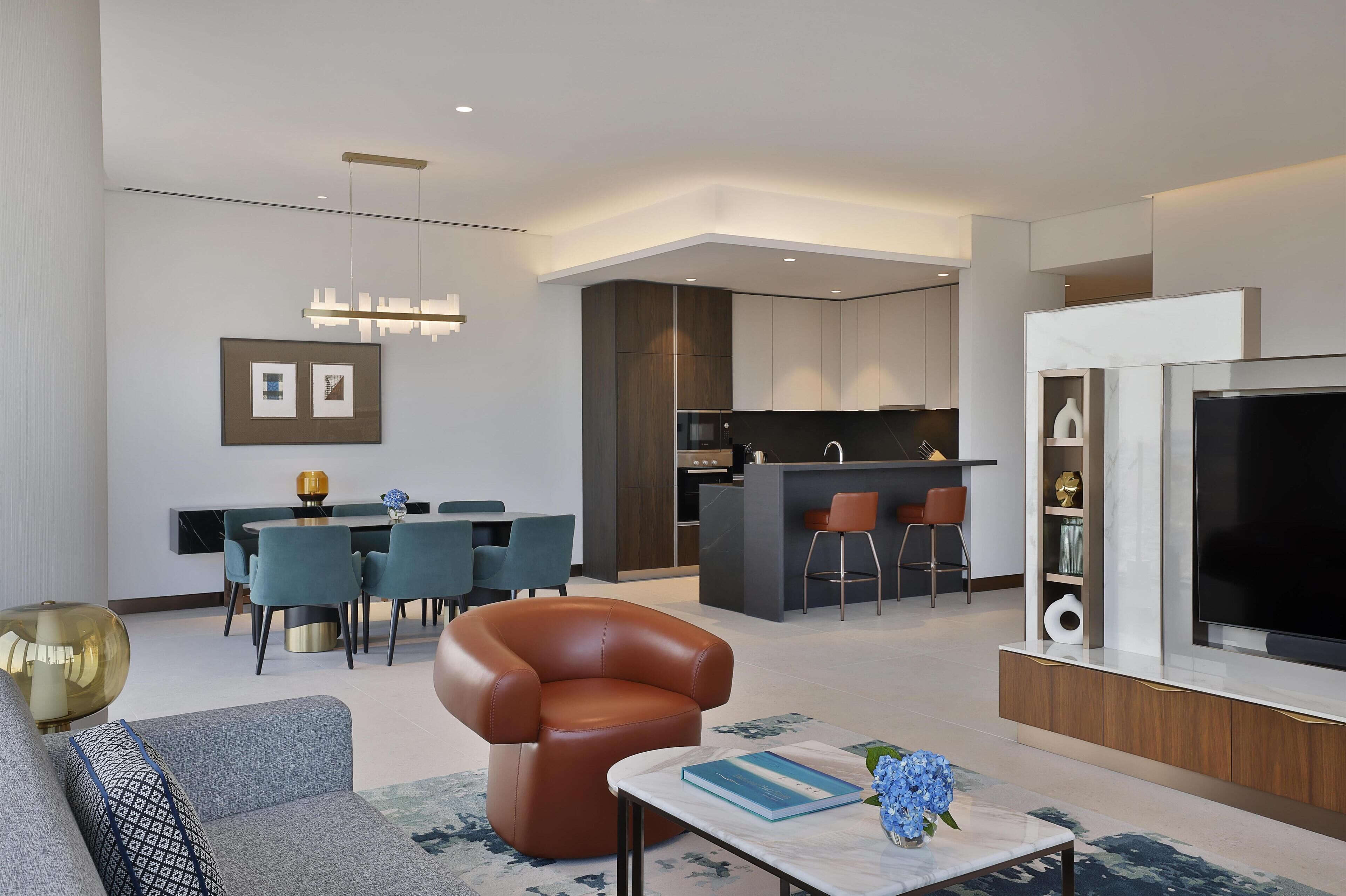 A modern living room with a sofa, coffee table, and a TV. There is a dining area with chairs and a table, and a kitchen with a bar.