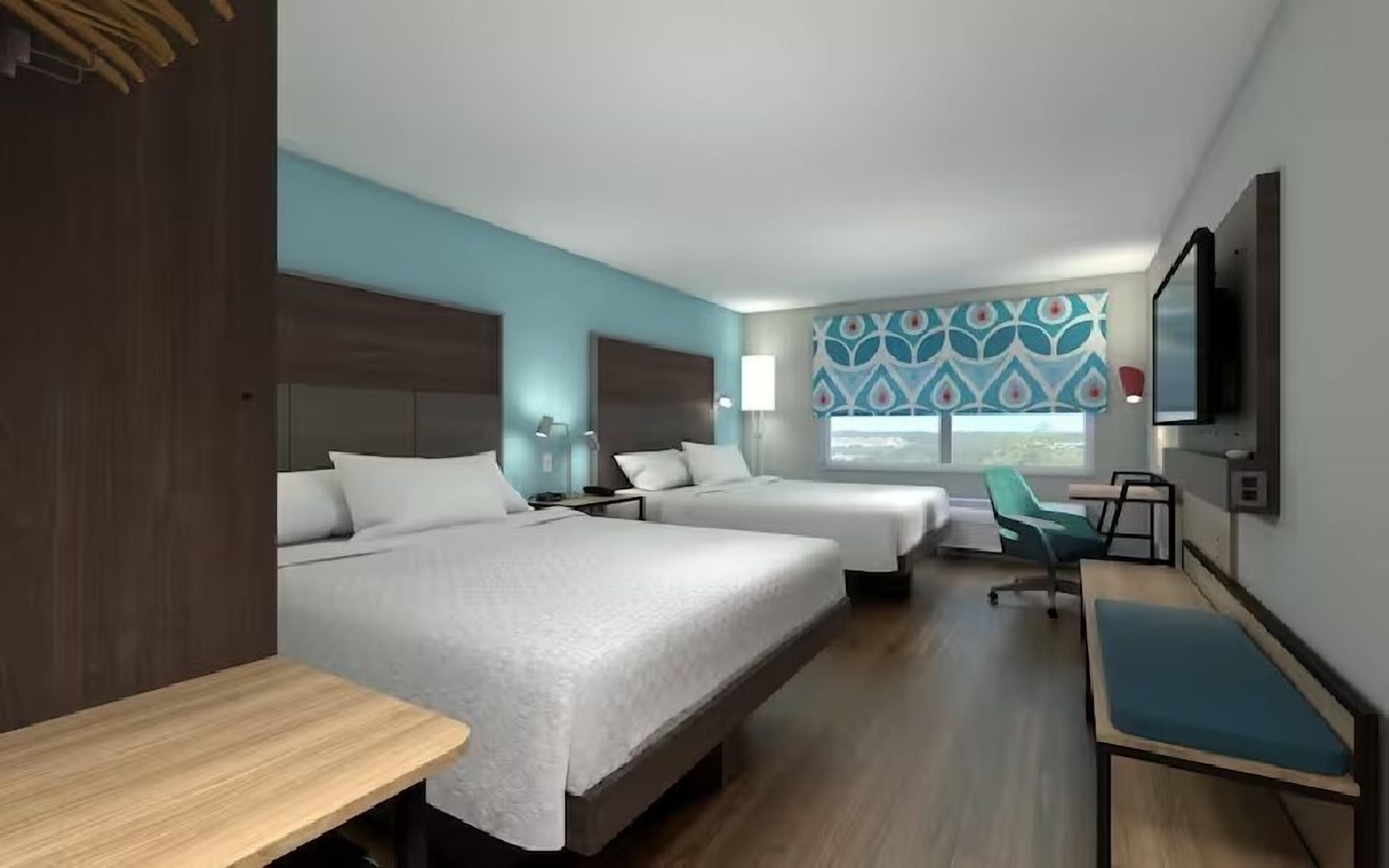 Habitación de hotel con dos camas, un escritorio, una silla, un televisor y una ventana con cortinas estampadas.