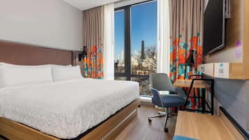 A hotel room with a large bed, a desk with a chair, a TV, and a view of the cityscape through the window.
