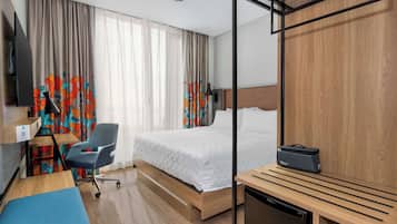 A hotel room with a bed, a desk with a chair, a small kitchenette, and a TV.