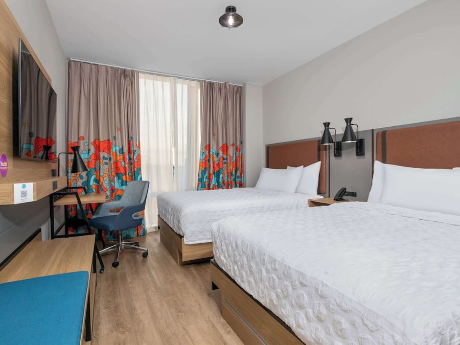 Quarto de hotel com duas camas, uma escrivaninha com cadeira, um espelho e uma janela com cortinas.