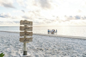 A wooden sign with the words "Quench Love Thirst Here" on a beach with people in the background. - Waldorf Astoria Seychelles Platte Island (Platte Island)