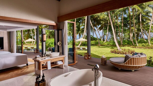 A modern bathroom with a freestanding bathtub, a wooden vanity, and a large window offering a view of a tropical garden and palm trees. - Waldorf Astoria Seychelles Platte Island (Platte Island)