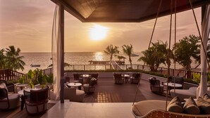 A covered outdoor seating area with tables and chairs, overlooking the sea at sunset.