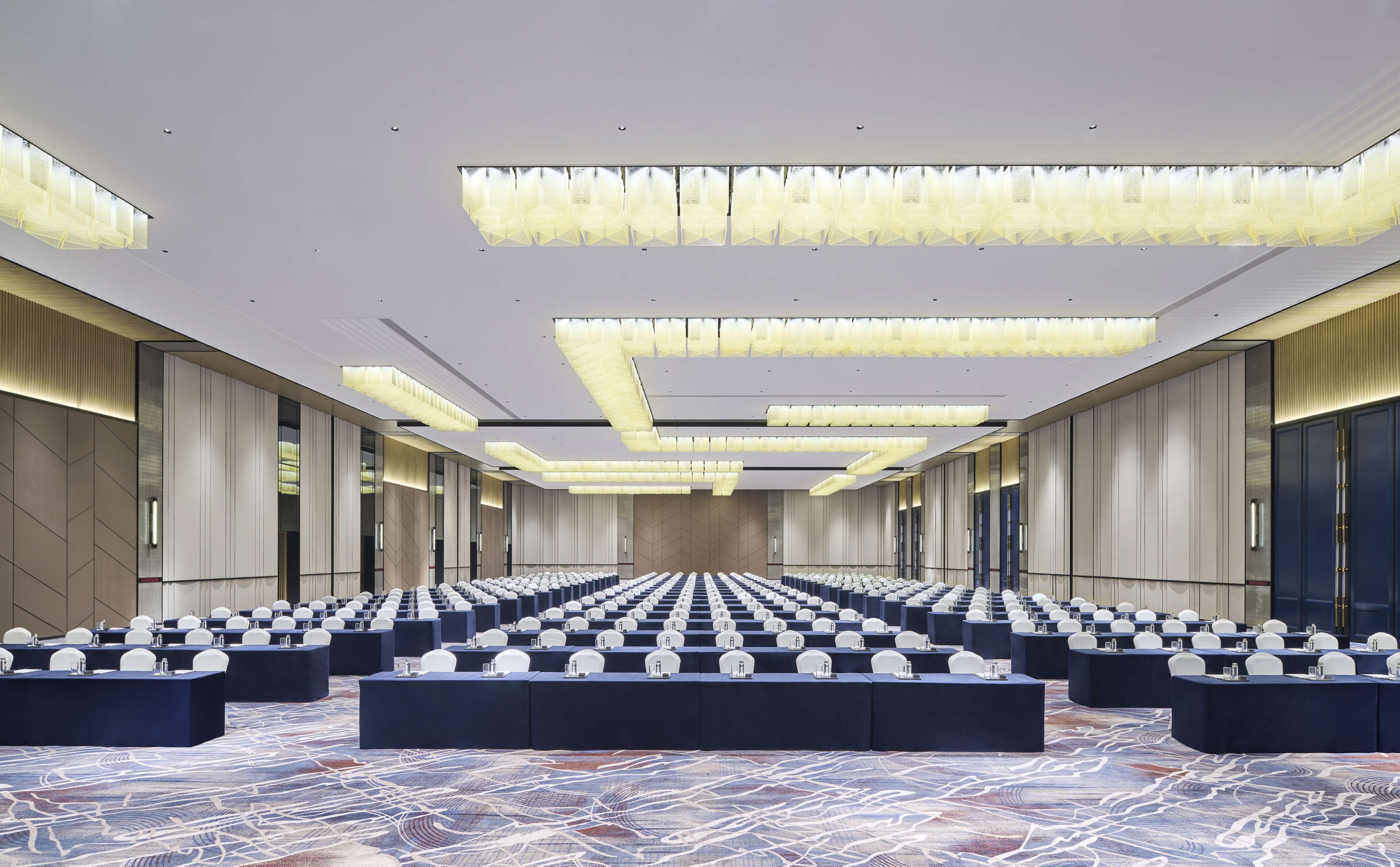 A large conference room with rows of tables and chairs, modern lighting fixtures, and a patterned carpet.