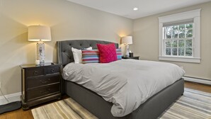 A bedroom with a large bed, two bedside tables with lamps, a window with a view of trees, and a striped rug. - Brook Bound Inn - Main Lodge - Deluxe King w/ Pullout Couch - Room 203
 (Wilmington)