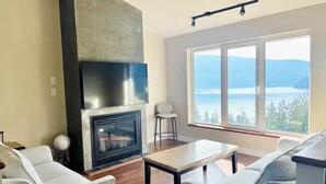 A modern living room with a fireplace, a large window with a view of a lake and mountains, and a dining area with a table and chairs.