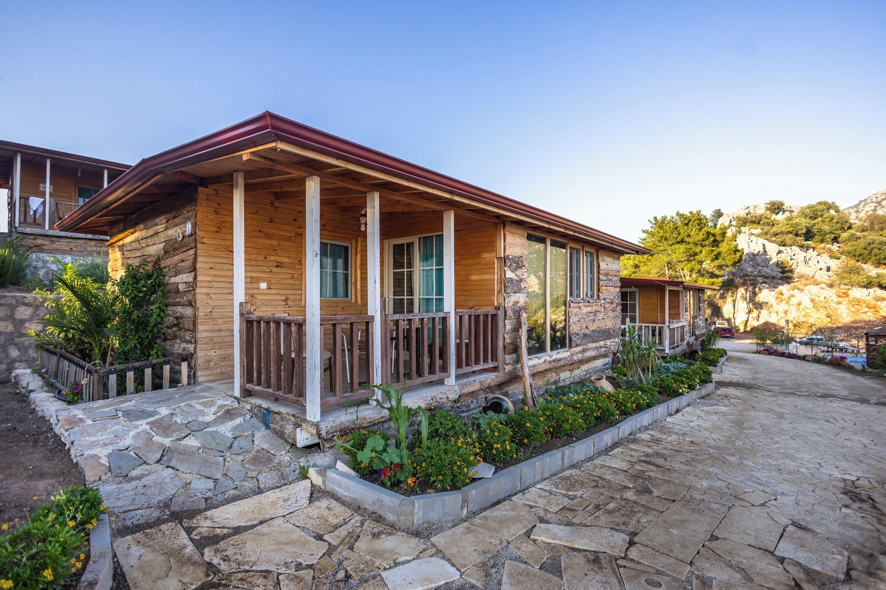 A wooden cabin with a stone pathway leading to the entrance, surrounded by greenery and a stone wall.