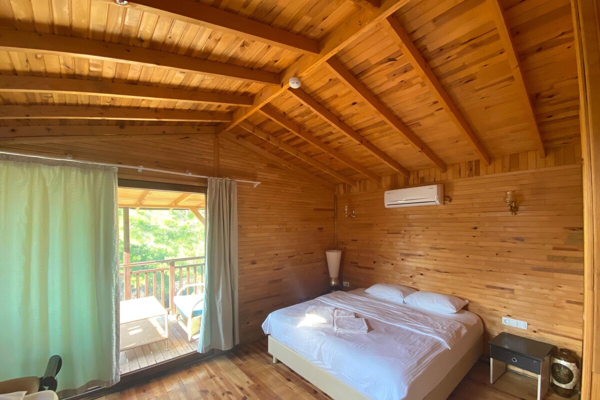 A wooden cabin bedroom with a bed, a nightstand, an air conditioning unit, and a view of the outdoors.
