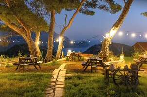 An outdoor dining area with picnic tables and benches, surrounded by trees and overlooking a body of water.