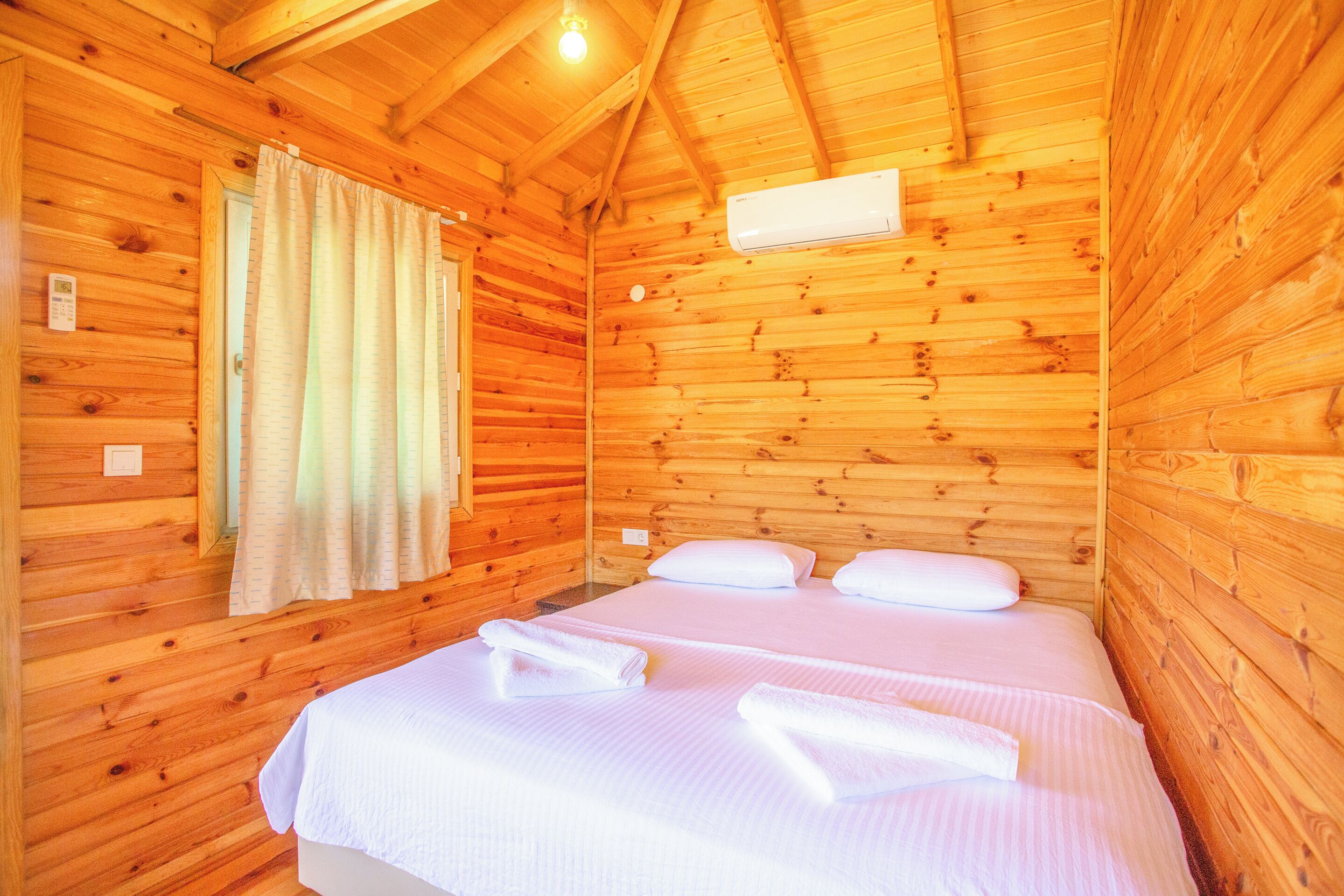 Une chambre dans un chalet en bois, comprenant un lit, deux oreillers, une fenêtre avec des rideaux et un appareil de climatisation.