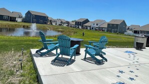 En patio med Adirondack-stole og et bålsted, en sø med en springvand, og en række byhuse i baggrunden.