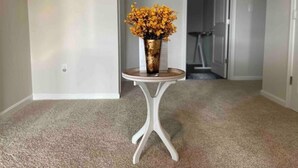 En gang med tæppe, en hvid væg med en lille hylde, og en vase med gule blomster på et hvidt piedestalbord.